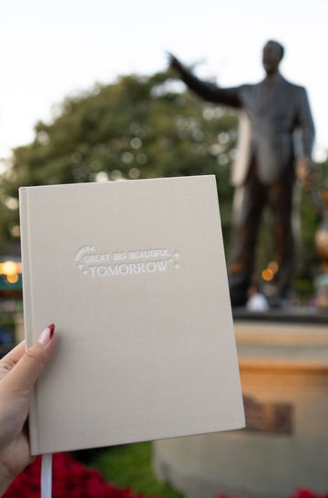 a neutral linen planner that says there's a great big beautiful tomorrow in white embossed text being held in front of the partner statue of walt and mickey at disneyland