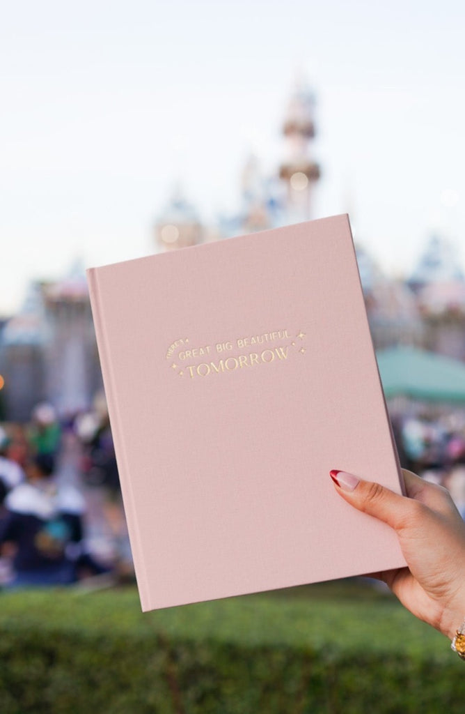 Pink fabric covered planner with gold foil that says there's a great big beautiful tomorrow being held up in front of sleeping beauty's castle at Disneyland